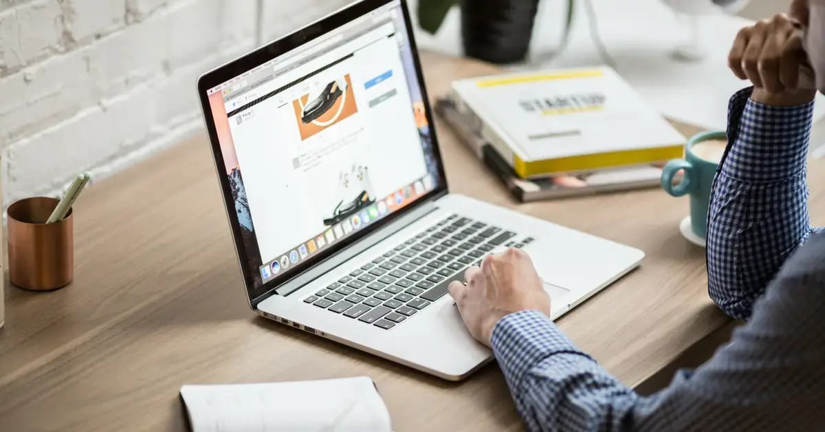 A person works on a laptop at a wooden desk, with a coffee mug and a book about startups to the side